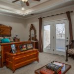 A traditional bedroom with a wooden bed, matching furniture, framed photos on a chest, a patterned chair, bookshelves, and French doors leading outside. Neutral walls and carpet, with a ceiling fan and decorative lighting.