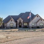 A large, single-story brick house with arched windows and a steep gabled roof sits behind a lawn and a flagpole, surrounded by trees and open land under a clear sky.