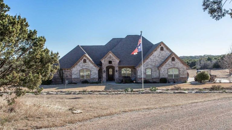 A large, single-story brick house with arched windows and a steep gabled roof sits behind a lawn and a flagpole, surrounded by trees and open land under a clear sky.