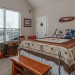 A cozy bedroom with a large window, bed with a brown and beige quilt, wooden bench, vintage trunk, chairs, nightstands with lamps, and stuffed animals displayed on the bed. Light walls and carpeted floor.