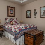 A cozy bedroom with a bed covered in a star-patterned quilt, stuffed animals, a wooden chest at the foot, two side tables with lamps, and nature-themed artwork on tan walls. Sunlight streams through a window with blinds.