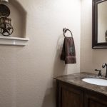 A bathroom with a dark wood vanity, marble countertop, oval sink, and black faucet. A brown towel hangs on the wall, and a shelf displays decor above the toilet. A large mirror is mounted above the sink.