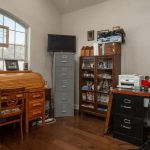 A home office with a wooden roll-top desk, chair, tall filing cabinet, bookshelf with office supplies, framed sports photos on the wall, and a printer on a black desk. Sunlight shines through a large window.