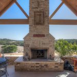 Outdoor covered patio with a stone fireplace, round black metal table and chairs, wooden ceiling, and scenic view of trees and open landscape in the background under a clear sky.