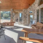 Spacious covered patio with brick walls, wooden picnic tables, a rocking chair, a metal table with chairs, a fireplace, and large windows overlooking a yard and trees.