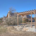 A wooden pergola stands over a circular arrangement of large rocks in a dry, grassy yard with a stone house and trees in the background under a clear blue sky.