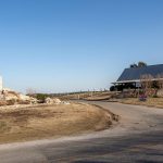 A red and white striped lighthouse stands on a stone base near a country road, with a wooden cabin and trees in the background under a clear blue sky.