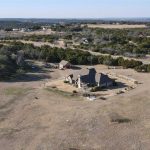 Aerial view of a house with a steep roof surrounded by dry grassland, trees, and a few buildings in the distance under a clear sky in a rural landscape.