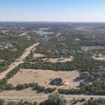 Aerial view of a large house surrounded by open land and dense trees, with scattered homes and winding roads throughout a hilly rural landscape under a clear sky.