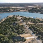 Aerial view of a peninsula with houses surrounded by trees, bordered by a river or lake on both sides, with rolling hills and distant water visible under a clear sky.