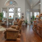 Spacious living room with wooden floors, brown recliner chairs, an ornate wooden cabinet, a stone fireplace, large windows, and decor items including lamps, plants, and framed pictures.