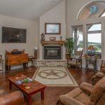 A cozy living room with leather chairs, a central fireplace, a wall-mounted TV, wooden furniture, and large arched windows letting in natural light. A patterned rug lies on the hardwood floor.