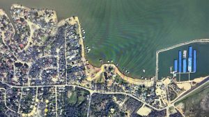 Aerial view of a lakeside residential area with houses, streets, docks extending into the water, and a marina with blue-roofed boat slips along the shoreline. Dense trees and curved roads surround the lake.
