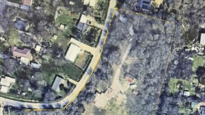 Aerial view of a residential area with houses, trees, and a curved road; a yellow outline marks the boundary of a large property containing multiple structures and wooded areas.