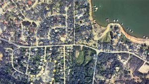Satellite view of a suburban neighborhood with curved and straight streets, houses, trees, and docks along the edge of a body of water at the top right. Yellow dots highlight a section of the area.