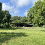 A grassy field with scattered trees under a blue sky with clouds. In the background, there are a few small, open-sided sheds or shelters surrounded by greenery.