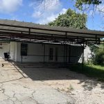 A metal carport with a corrugated roof stands on a cracked concrete driveway in front of a white house, surrounded by green trees and grass under a partly cloudy sky.