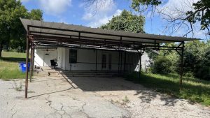 A metal carport with a corrugated roof stands on a cracked concrete driveway in front of a white house, surrounded by green trees and grass under a partly cloudy sky.