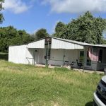 A small white house with a covered porch sits in a grassy yard under a partly cloudy sky. An American flag hangs by the porch, and a white vehicle is parked in the foreground. Trees surround the home.