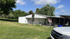 A small white house with a covered porch sits in a grassy yard under a partly cloudy sky. An American flag hangs by the porch, and a white vehicle is parked in the foreground. Trees surround the home.