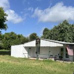 A white metal shed with a slanted roof sits in a grassy yard under a blue sky with clouds. An American flag hangs near the entrance, and trees surround the area. The edge of a white vehicle is visible in the foreground.