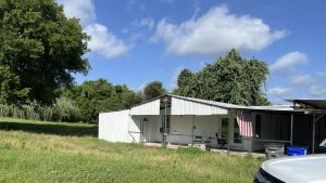 A white metal shed with a slanted roof sits in a grassy yard under a blue sky with clouds. An American flag hangs near the entrance, and trees surround the area. The edge of a white vehicle is visible in the foreground.