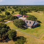Aerial view of a large brick house with a fenced yard, surrounded by trees and open fields. Nearby are two red-roofed outbuildings and a driveway leading to the property, set in a rural landscape.