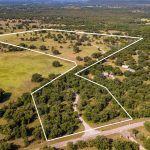 Aerial view of a rural landscape with a large, irregularly shaped plot of land outlined in white, featuring open fields, scattered trees, several homes, and a winding dirt road.