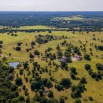 Aerial view of a large rural landscape with green fields, scattered trees, a pond, and a few buildings, stretching into the distance under a clear sky.