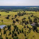 Aerial view of a rural landscape with scattered trees, a pond, a winding road, a house, and a red-roofed barn surrounded by open grassy fields under a clear sky.