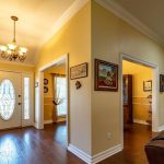 A warm, well-lit home entryway with a glass front door, chandelier, wooden floor, and several open doorways leading to other rooms. Wall art and a grandfather clock accent the hallway.