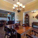 A dining room with a wooden table and chairs under a chandelier, leading into a living room with sofas and a fireplace. An antique grandfather clock stands in the hallway to the right. Walls are light yellow with white trim.