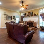 A spacious living room with hardwood floors, brown leather sofas, a brick fireplace with a white mantel, wall art, and large windows with plaid curtains. Adjoining rooms and a ceiling fan are also visible.