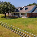 A large brick house with a covered front porch sits on a spacious grassy yard, bordered by a metal fence and dotted with lamp posts. A large tree provides shade near the house.