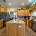 Spacious kitchen with wooden cabinets, granite countertops, a central island, stainless steel appliances, and decorative greenery above the cabinets. There are windows above the sink letting in natural light.
