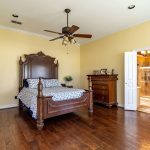 A spacious bedroom with wooden floors, a large wooden bed with blue and white bedding, a matching dresser, and ceiling fan. Double doors lead to a tiled bathroom, and another door is open to the left.