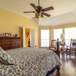 A spacious bedroom with yellow walls, a patterned bedspread, wooden floor, ceiling fan, dresser, and large windows letting in natural light. Two rocking chairs and a lamp sit by the windows.