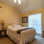A cozy bedroom with yellow walls, a double bed with white bedding, a bedside table with a lamp, artwork on the walls, a window with blinds, and a wooden chair in the corner.