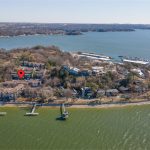 Aerial view of a lakeside neighborhood with docks extending into the water, houses surrounded by trees, and a red location pin marking one property near the shoreline. A marina with boats is visible in the background.