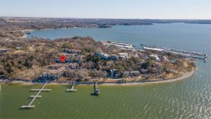 Aerial view of a lakeside neighborhood with docks extending into the water, houses surrounded by trees, and a red location pin marking one property near the shoreline. A marina with boats is visible in the background.