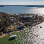 Aerial view of a peninsula with houses surrounded by water, a dock extending into the lake, and a red location marker above one house. The shoreline is lined with trees and the city skyline is visible in the distance.