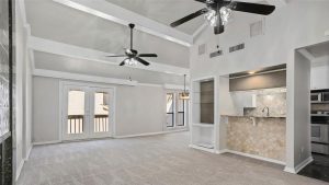Spacious living area with vaulted ceiling, two ceiling fans, French doors to a balcony, carpeted floor, and an open kitchen with a breakfast bar and tile backsplash. Dining area with large window is visible.