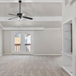 Spacious living room with beige carpet, light gray walls, a ceiling fan, and exposed beams. French doors lead to a balcony. An open kitchen with a breakfast bar is visible on the right.