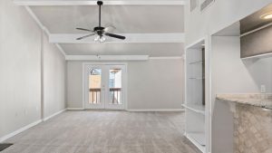 Spacious living room with beige carpet, light gray walls, a ceiling fan, and exposed beams. French doors lead to a balcony. An open kitchen with a breakfast bar is visible on the right.