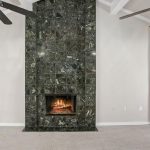 A room with beige carpet and light walls features a black marble-tiled fireplace with a lit fire. Two ceiling fans with lights and exposed beams are overhead. A wooden door is partially visible on the left.