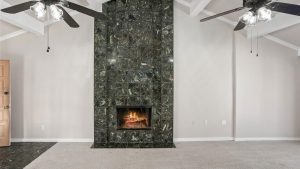 A room with beige carpet and light walls features a black marble-tiled fireplace with a lit fire. Two ceiling fans with lights and exposed beams are overhead. A wooden door is partially visible on the left.