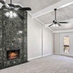 A modern living room with a tall, black marble fireplace, two ceiling fans with lights, light gray walls, carpeted floor, and double glass doors leading to a balcony.