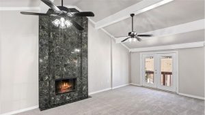 A modern living room with a tall, black marble fireplace, two ceiling fans with lights, light gray walls, carpeted floor, and double glass doors leading to a balcony.