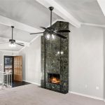 A spacious living room with vaulted ceilings, two ceiling fans, a dark marble fireplace with a fire burning, carpeted flooring, white walls, and large windows next to a wooden front door.