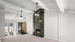 A spacious living room with vaulted ceilings, two ceiling fans, a dark marble fireplace with a fire burning, carpeted flooring, white walls, and large windows next to a wooden front door.